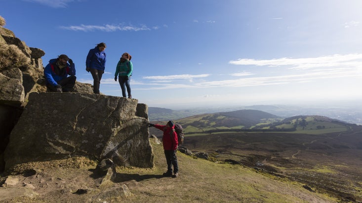 Walkers climbing rocks against a bright blue sky with the mountains in the distance at Sugarloaf, Monmouthshire